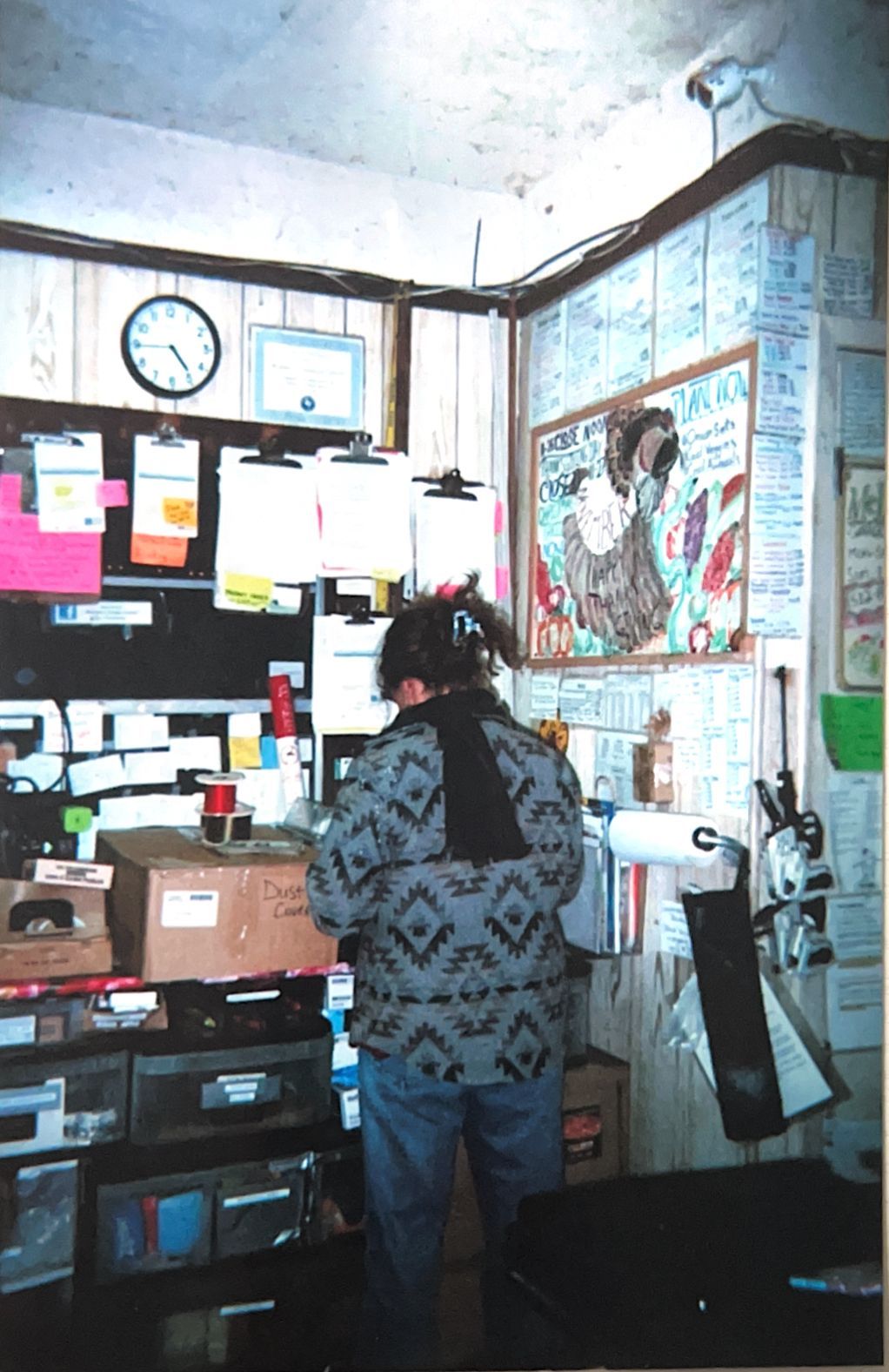 Person standing at a cluttered office desk with papers, boxes, and wall notices