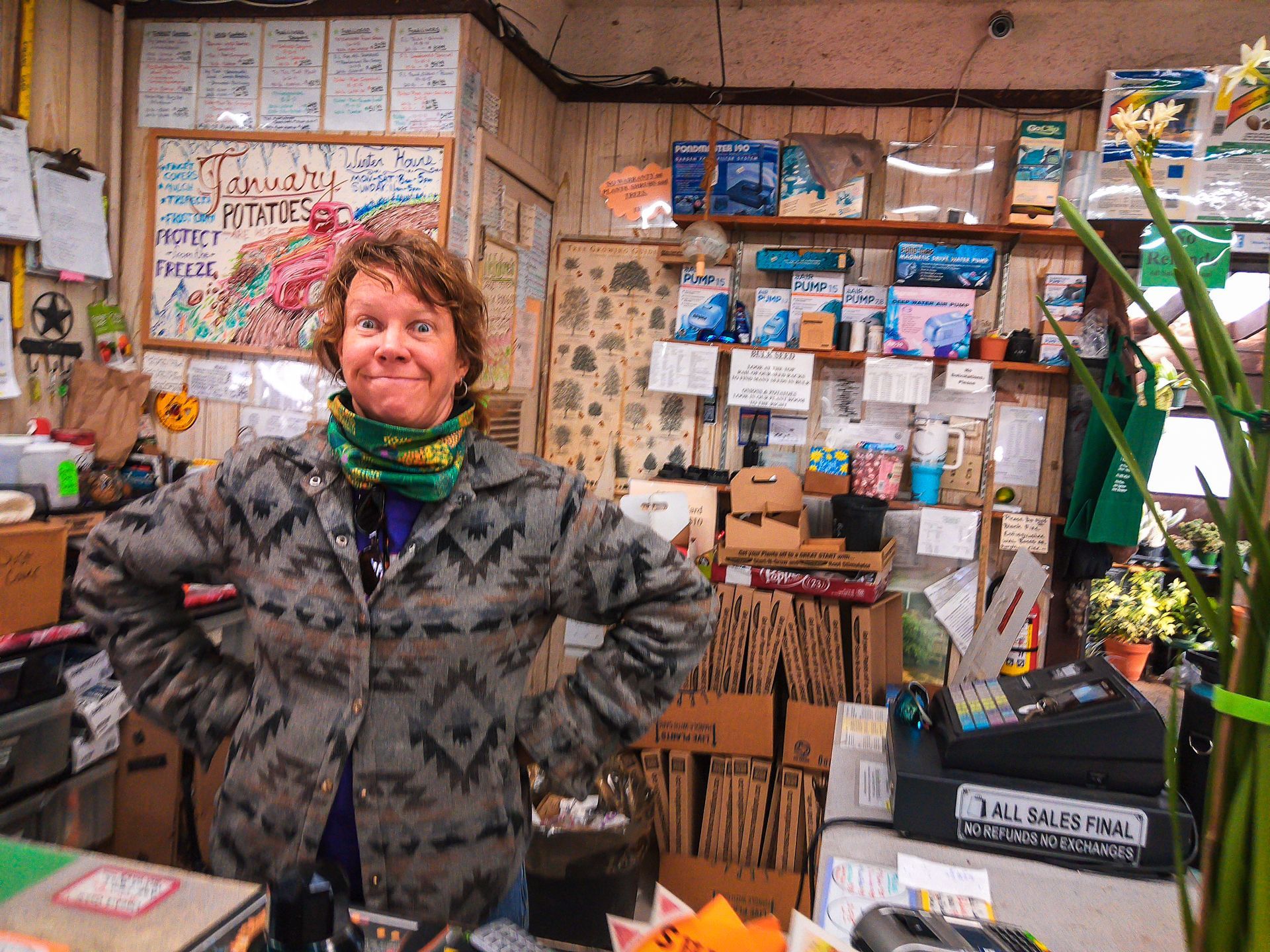 Person standing in a cluttered shop, smiling behind a counter amid shelves of small goods and signs.