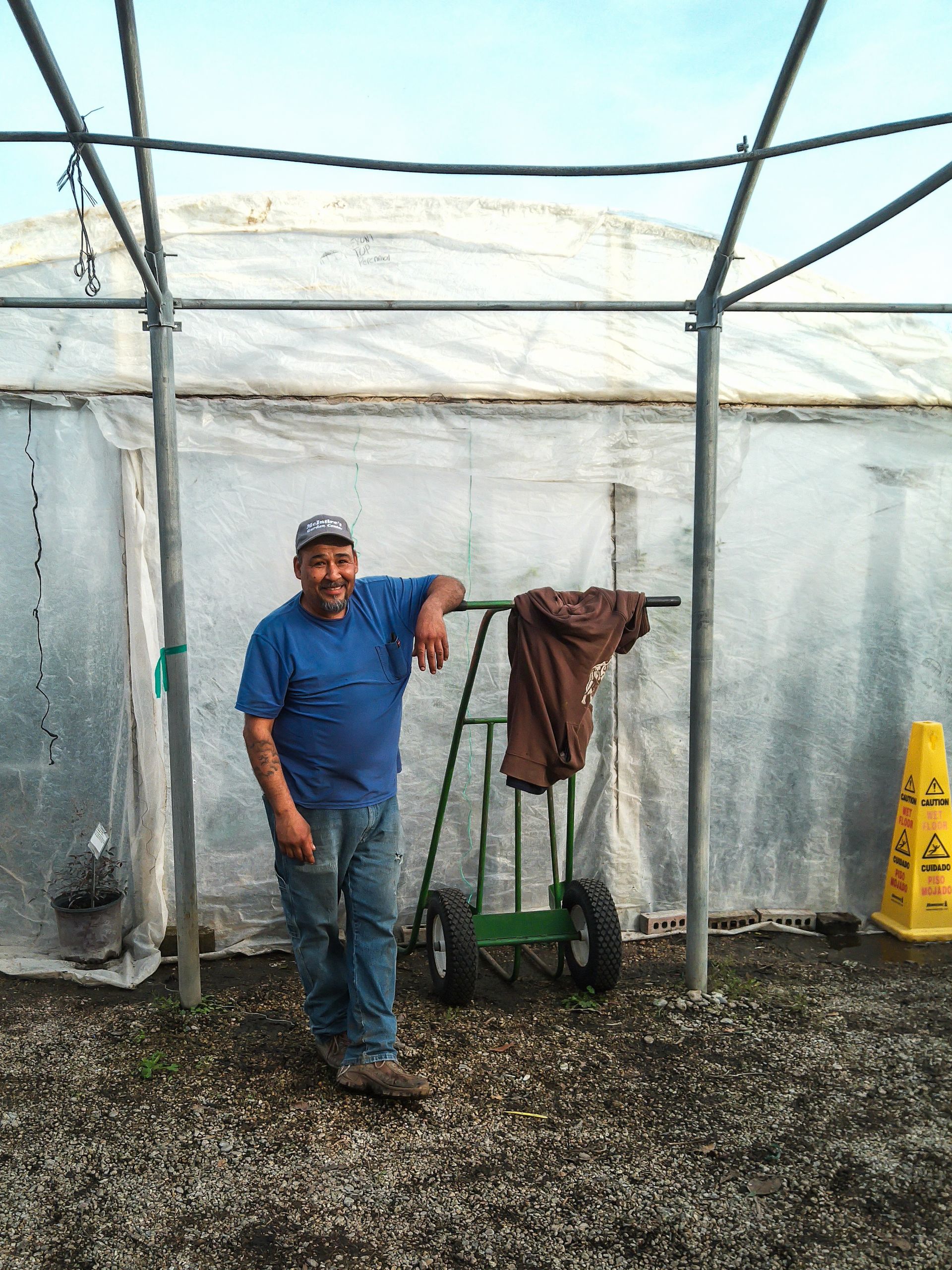 Man standing in a gravel enclosure beside a cart and hanging coat under a metal frame.