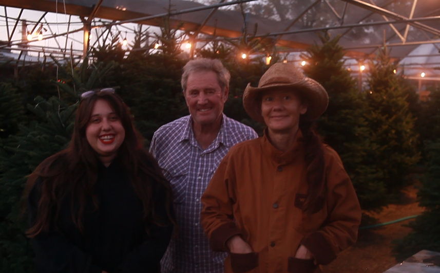Three people smiling in a greenhouse at dusk, with string lights and trees behind them.