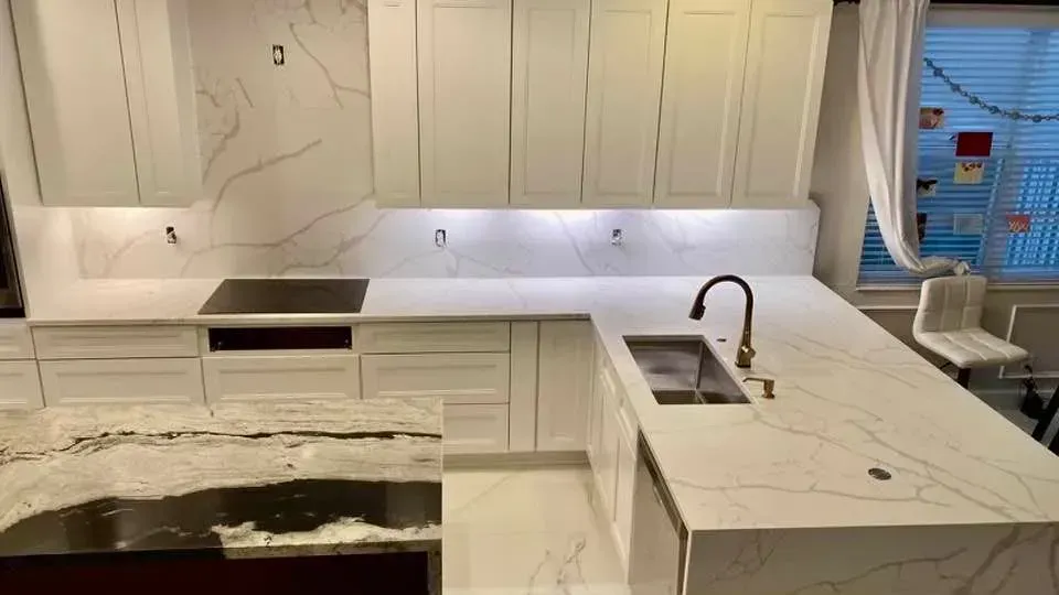 Modern white kitchen with marble countertops, sink, stovetop, and a window on the right