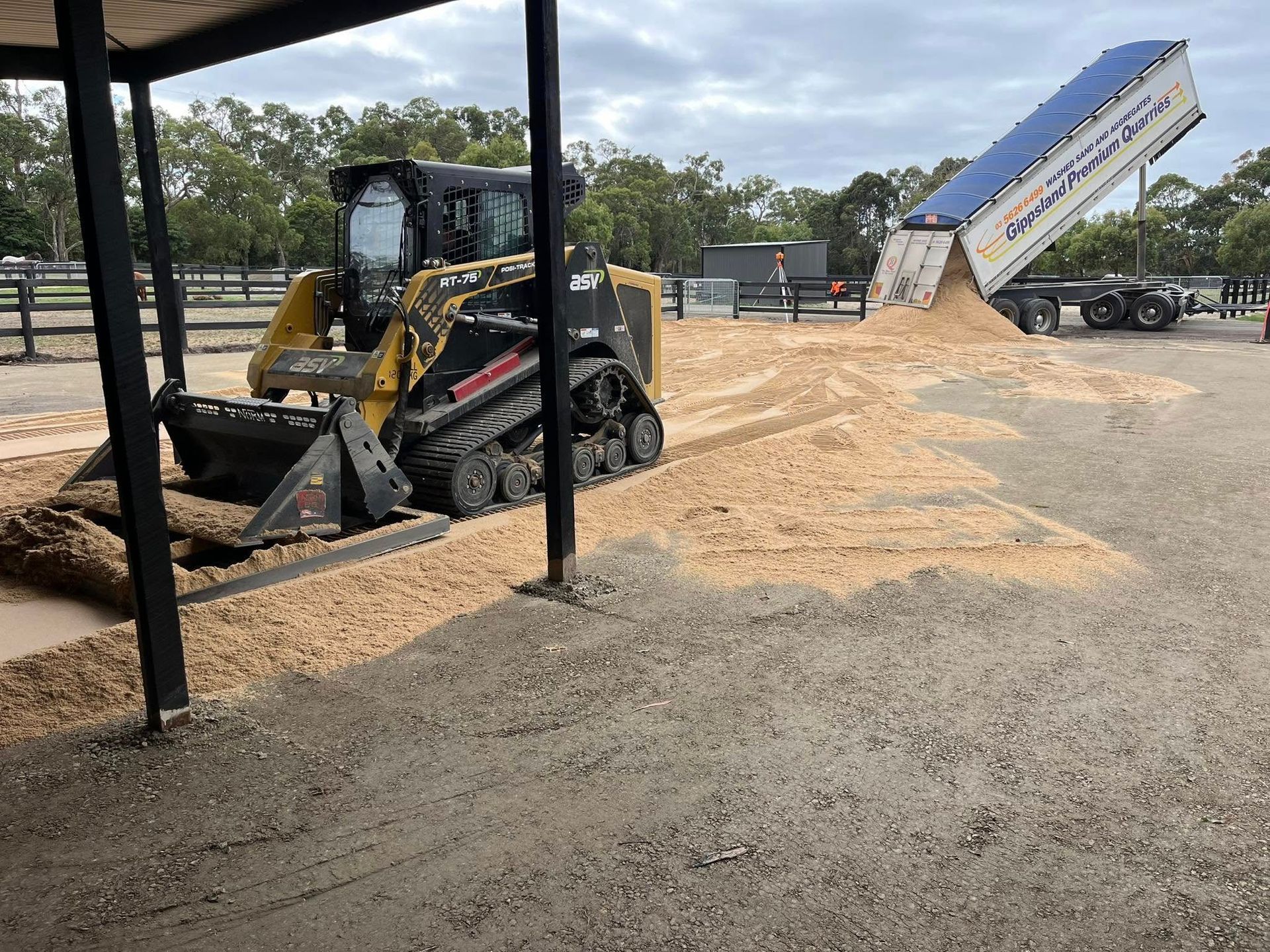 Skid steer loader moving wood chips under a shelter as a semi-trailer unloads more chips in an outdoor setting. — Equine Stable Solutions In Diddillibah, QLD