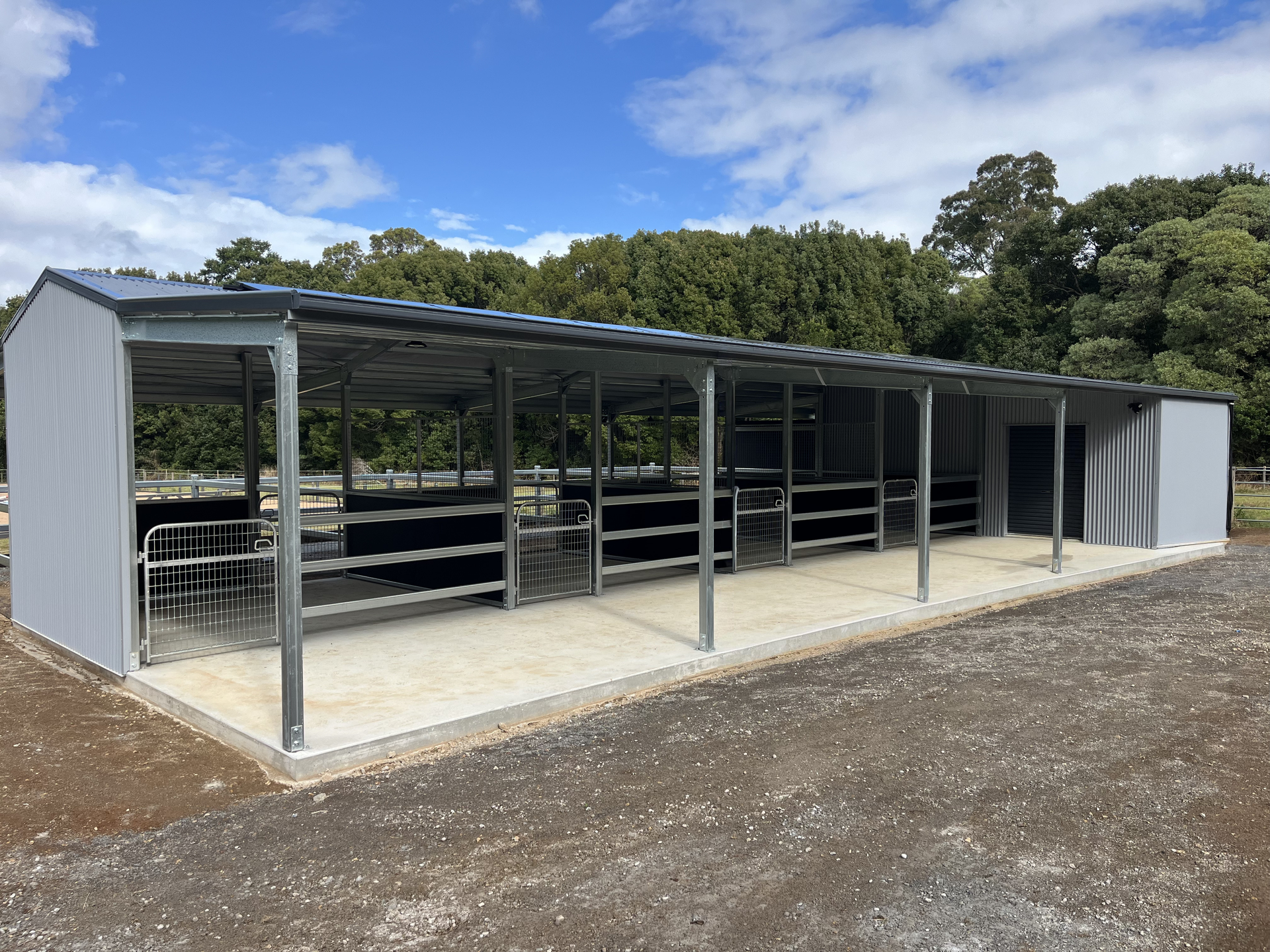 Gray metal farm building with stalls under a covered awning, blue sky background.