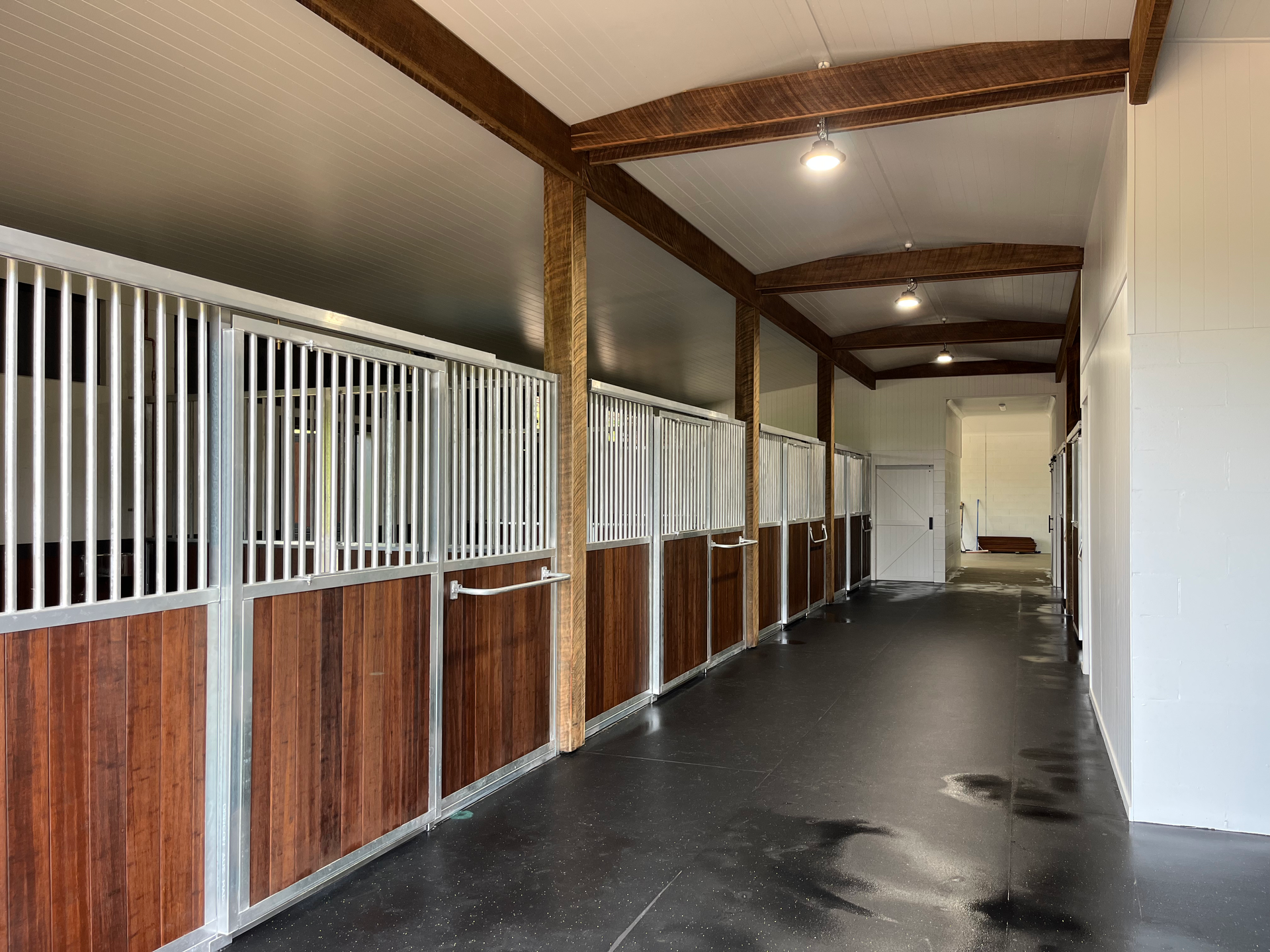 Horse stalls inside a stable with wooden doors and silver bars, a black floor, and a white ceiling — Equine Stable Solutions In Diddillibah, QLD