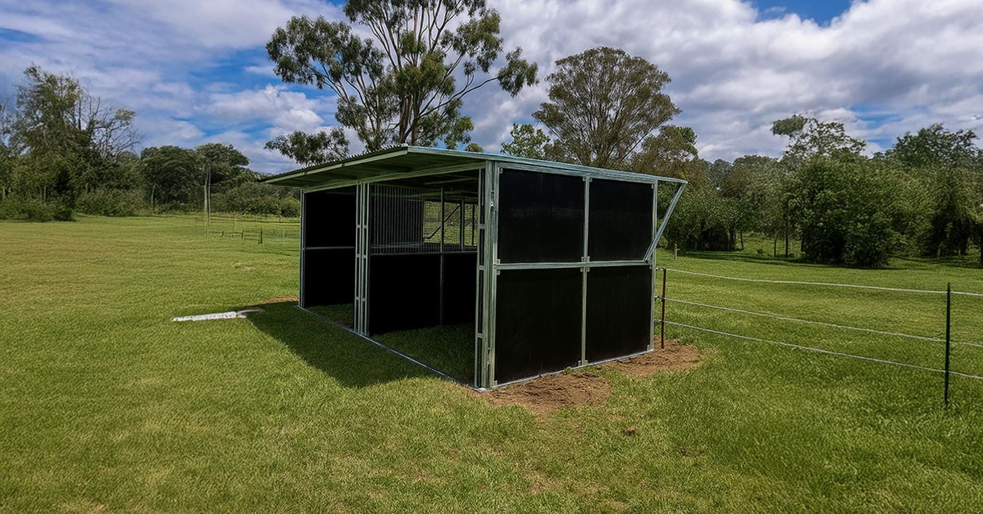 A metal horse shelter with a black interior stands on a grassy field under a blue sky.