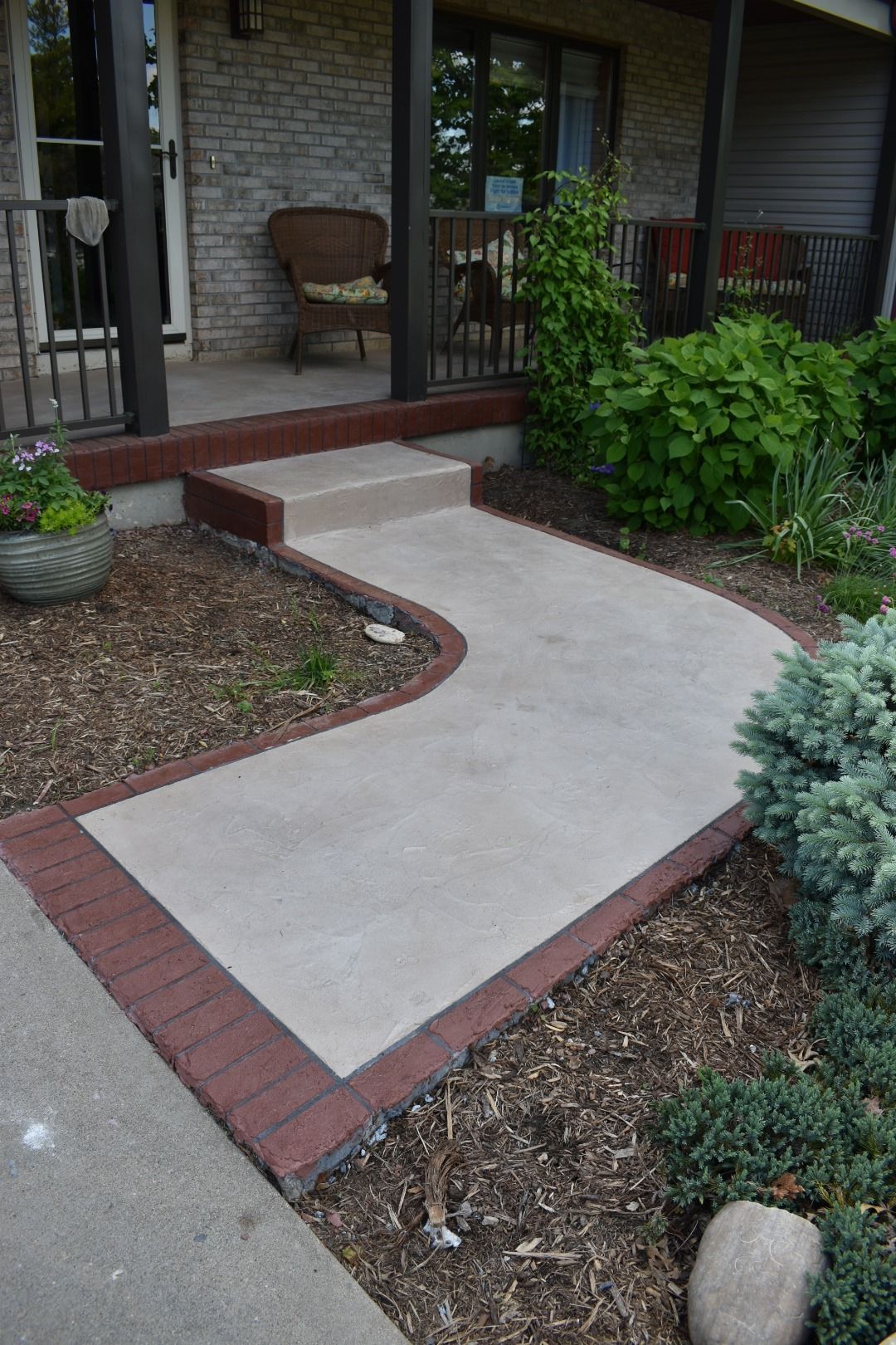 Curved concrete walkway with red brick edging leading to a front porch and garden plants
