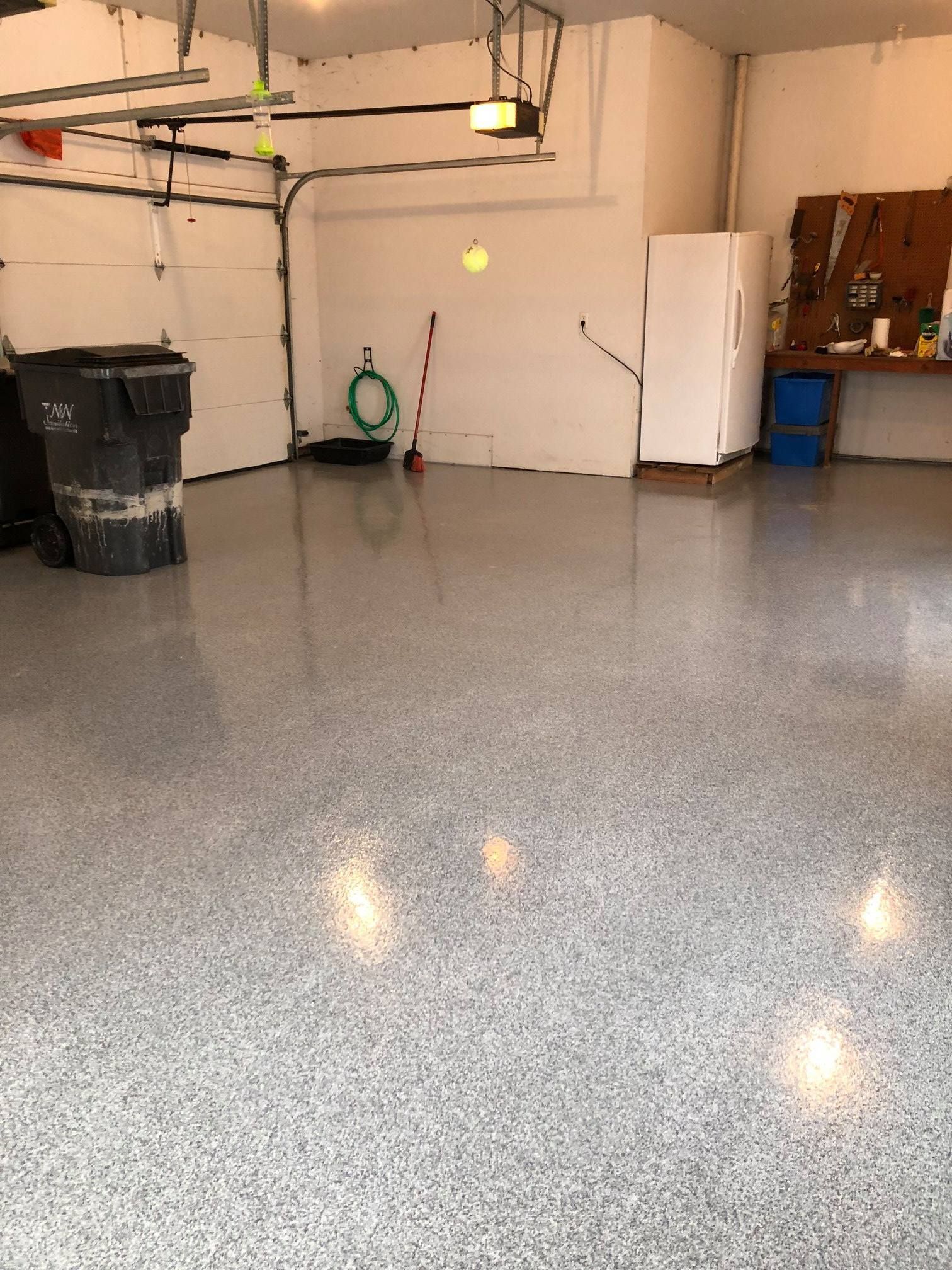 Empty garage with speckled epoxy floor, trash bin, storage shelves, and a white fridge against the back wall.