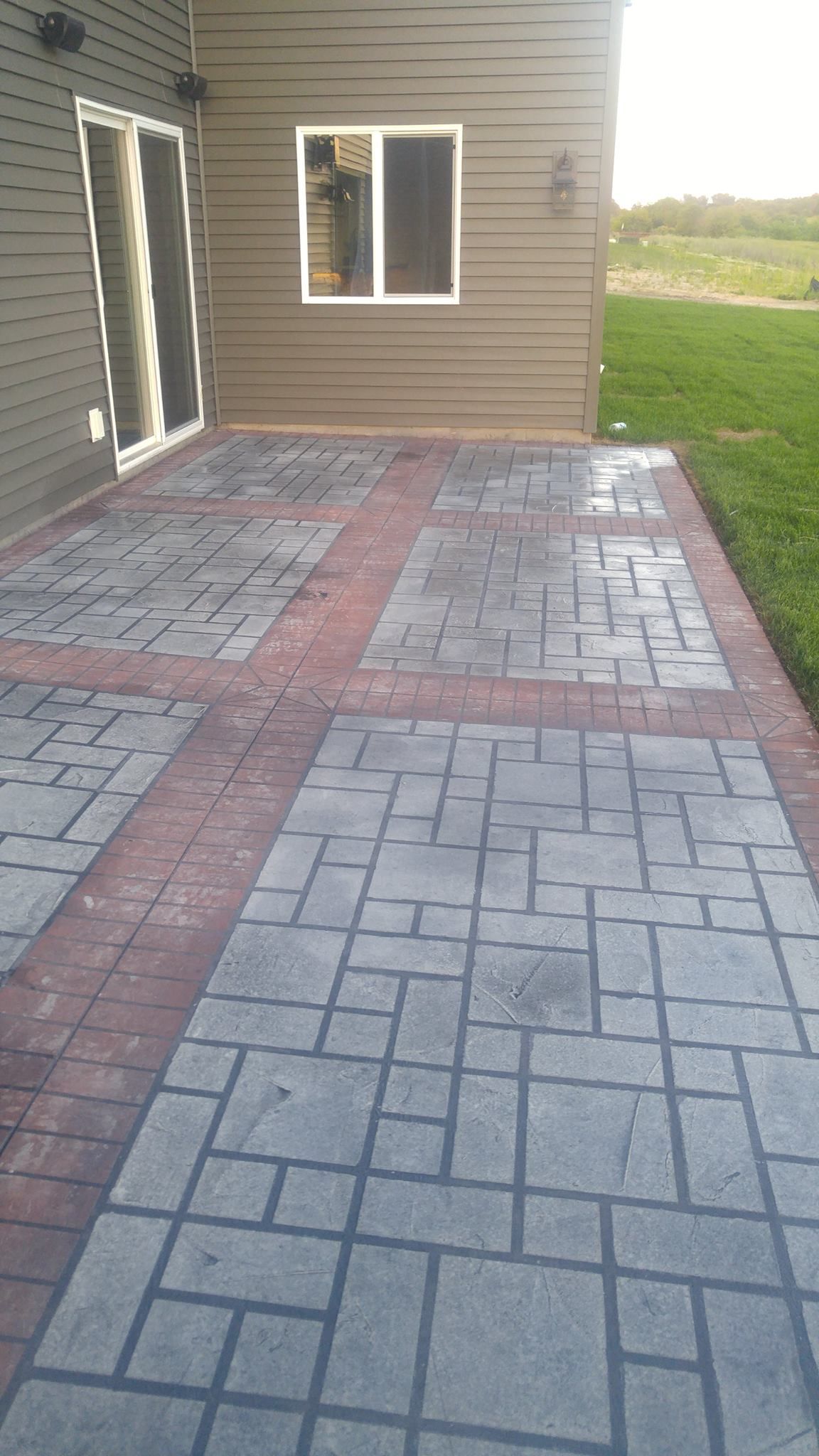 Patio with gray and red pavers beside a tan house, with a window and open door leading to a grassy yard
