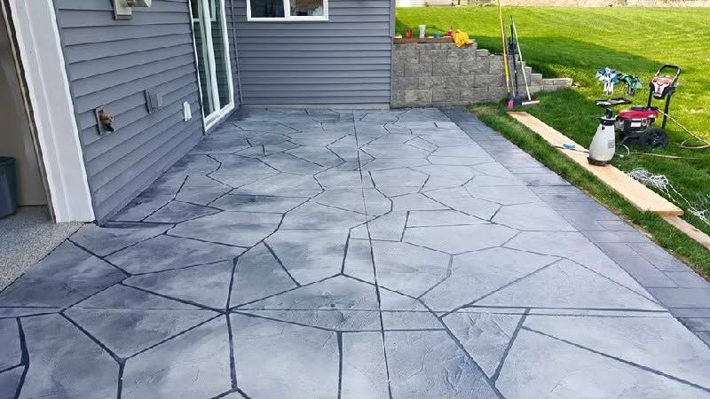 Stamped concrete patio beside a gray house, with patio tools and a grassy yard along the edge.