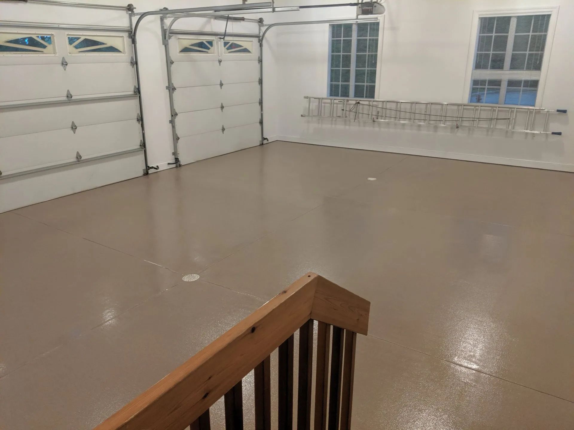 Empty garage with glossy floor, white walls, closed doors, windows, and a stair railing in the foreground