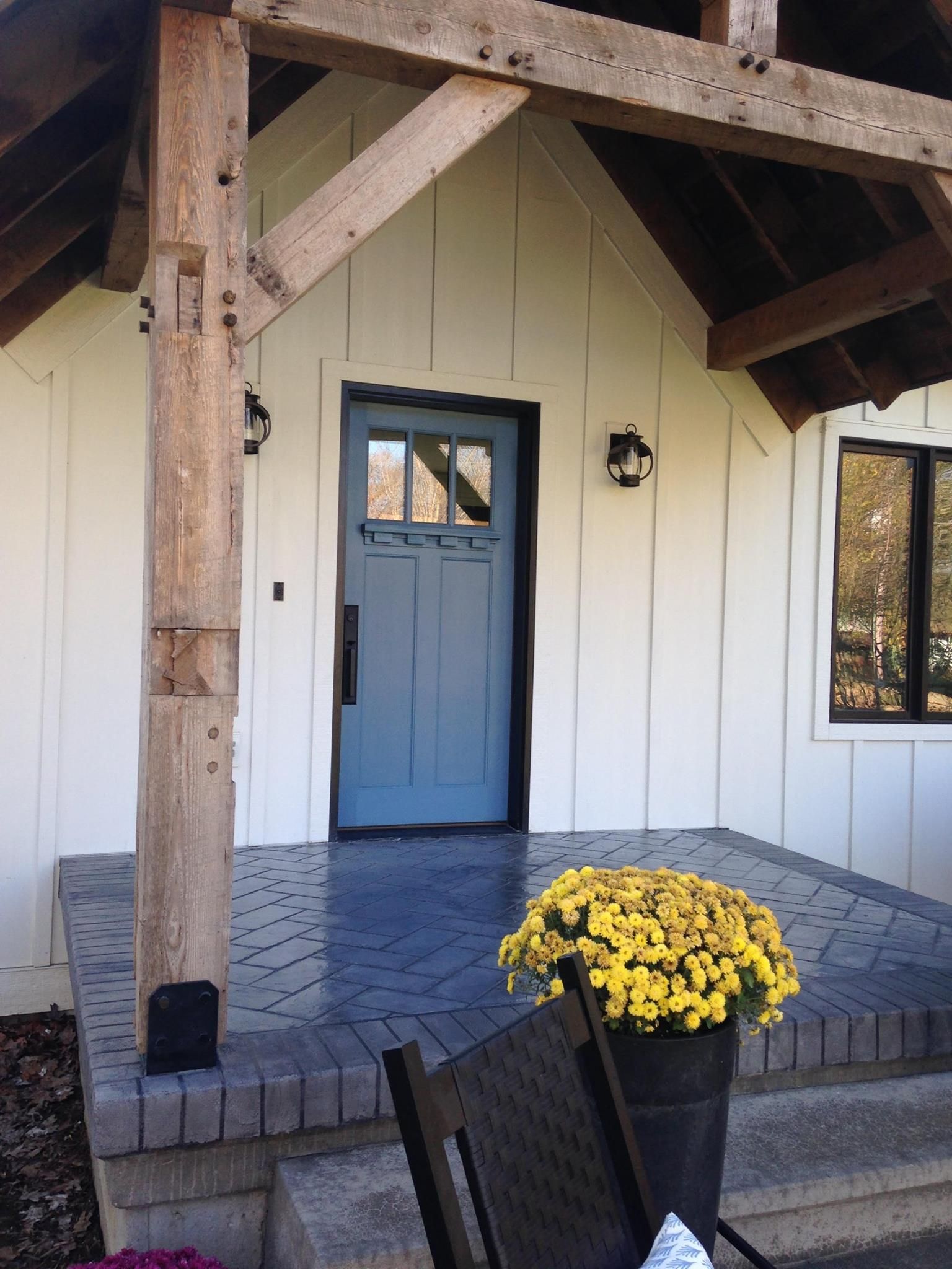 Front porch with a blue door, white siding, wooden beams, and a pot of yellow flowers on the steps