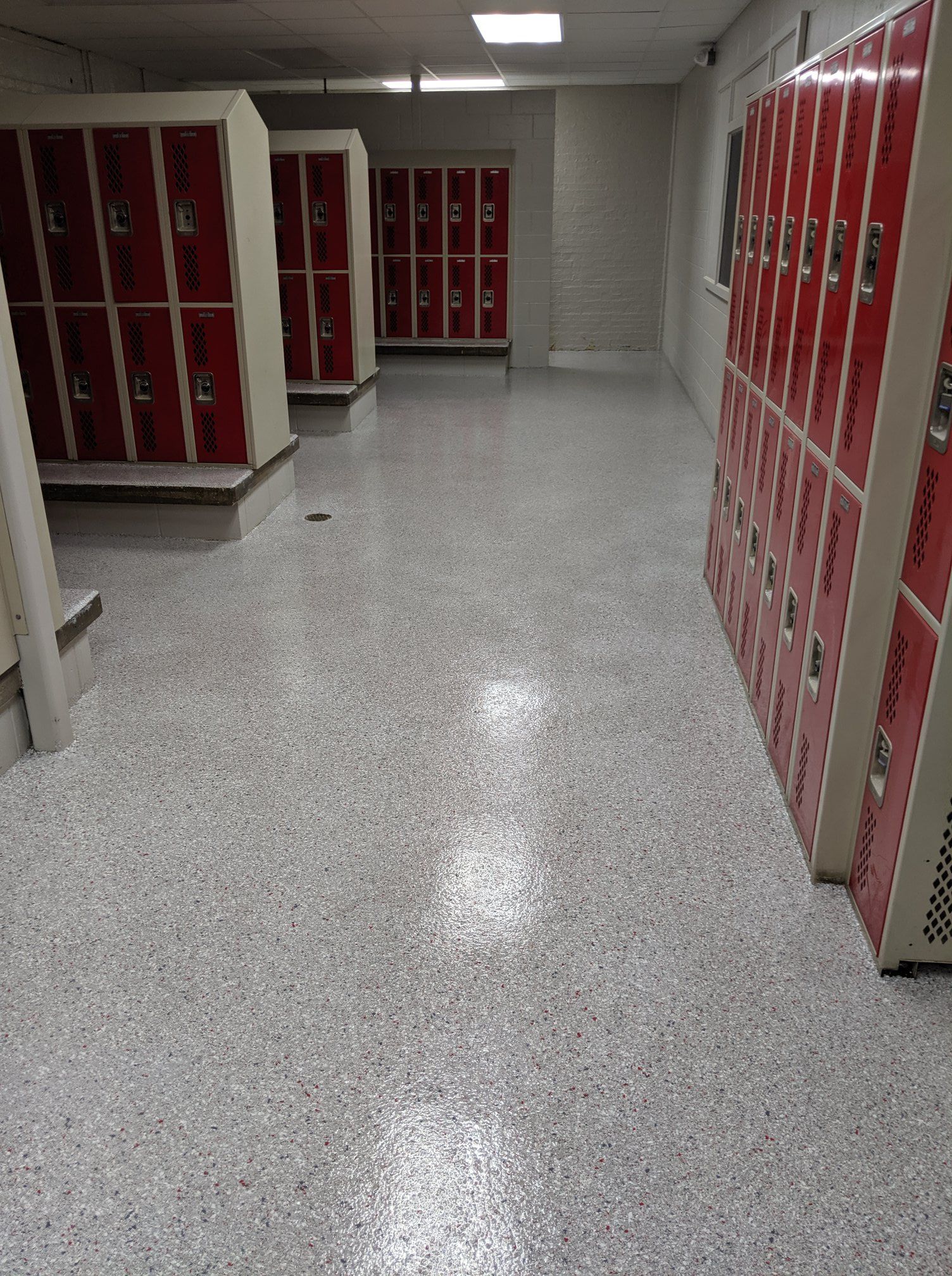 Quiet school hallway with red lockers lining both sides and a speckled gray floor.