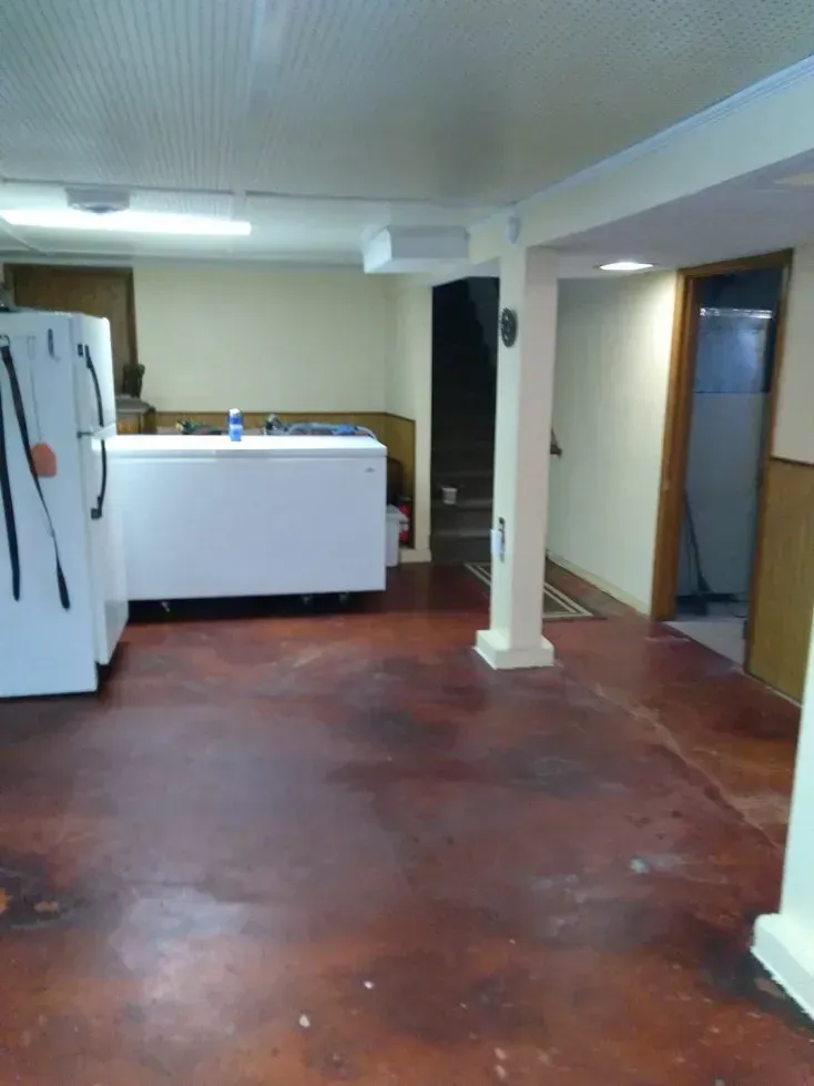 Bare basement room with a white chest freezer, fridge, and reddish-brown floor.