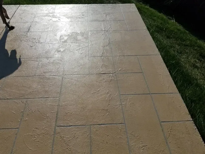 Wet tan stone patio beside a strip of green grass in sunlight