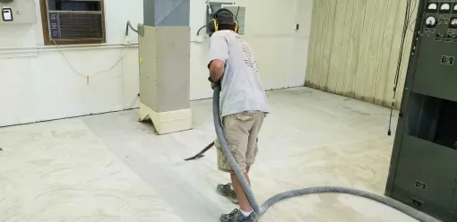 Worker operating a floor grinder in a bare indoor room with dust and equipment.