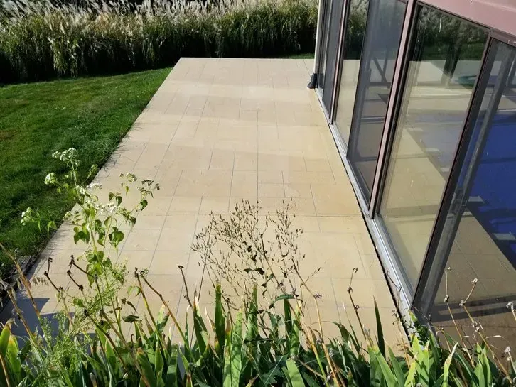 Patio with beige tiles beside a glass-walled house and green lawn bordering tall grass.