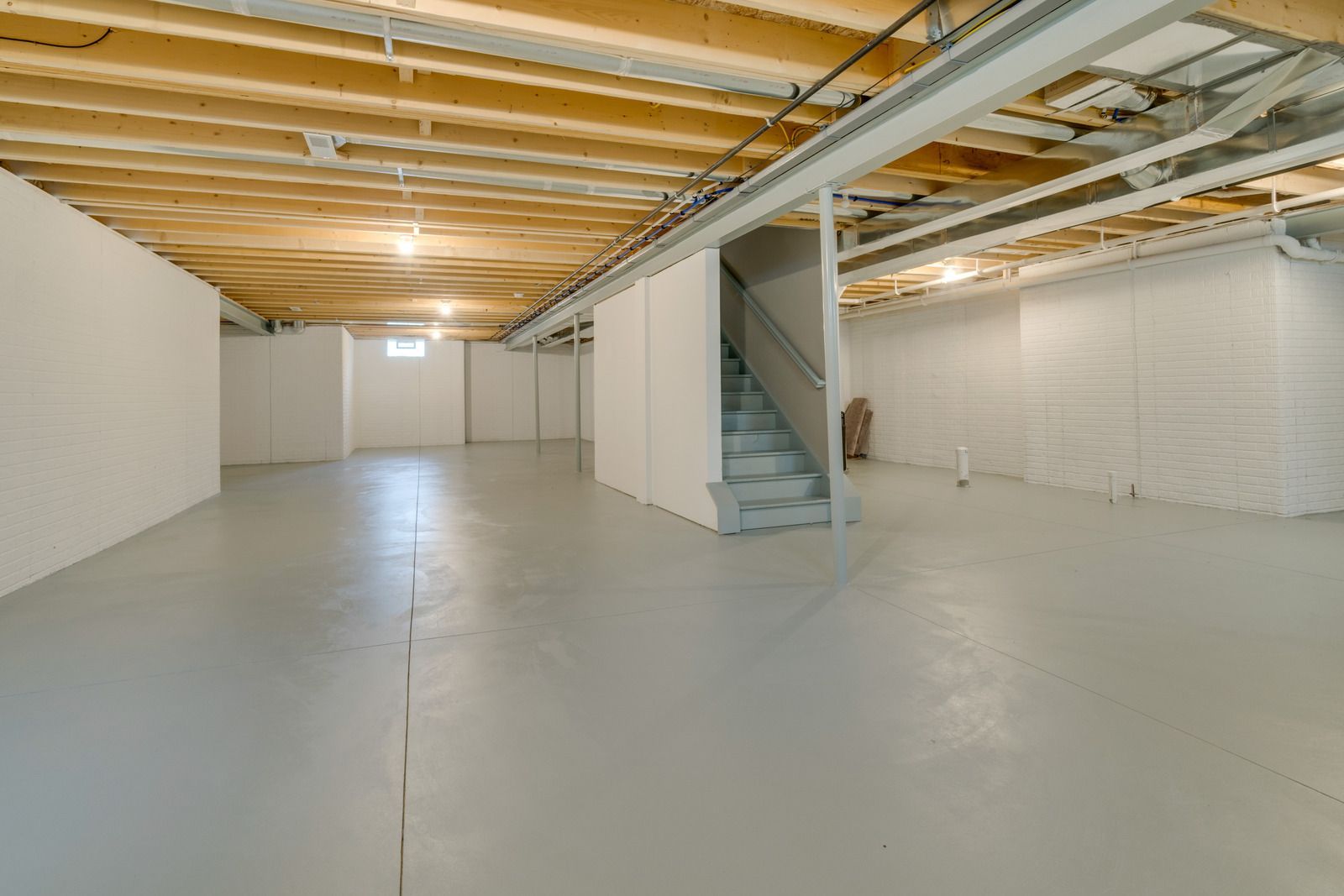 Empty basement with concrete floor, exposed wooden ceiling beams, white walls, and a staircase on the right.