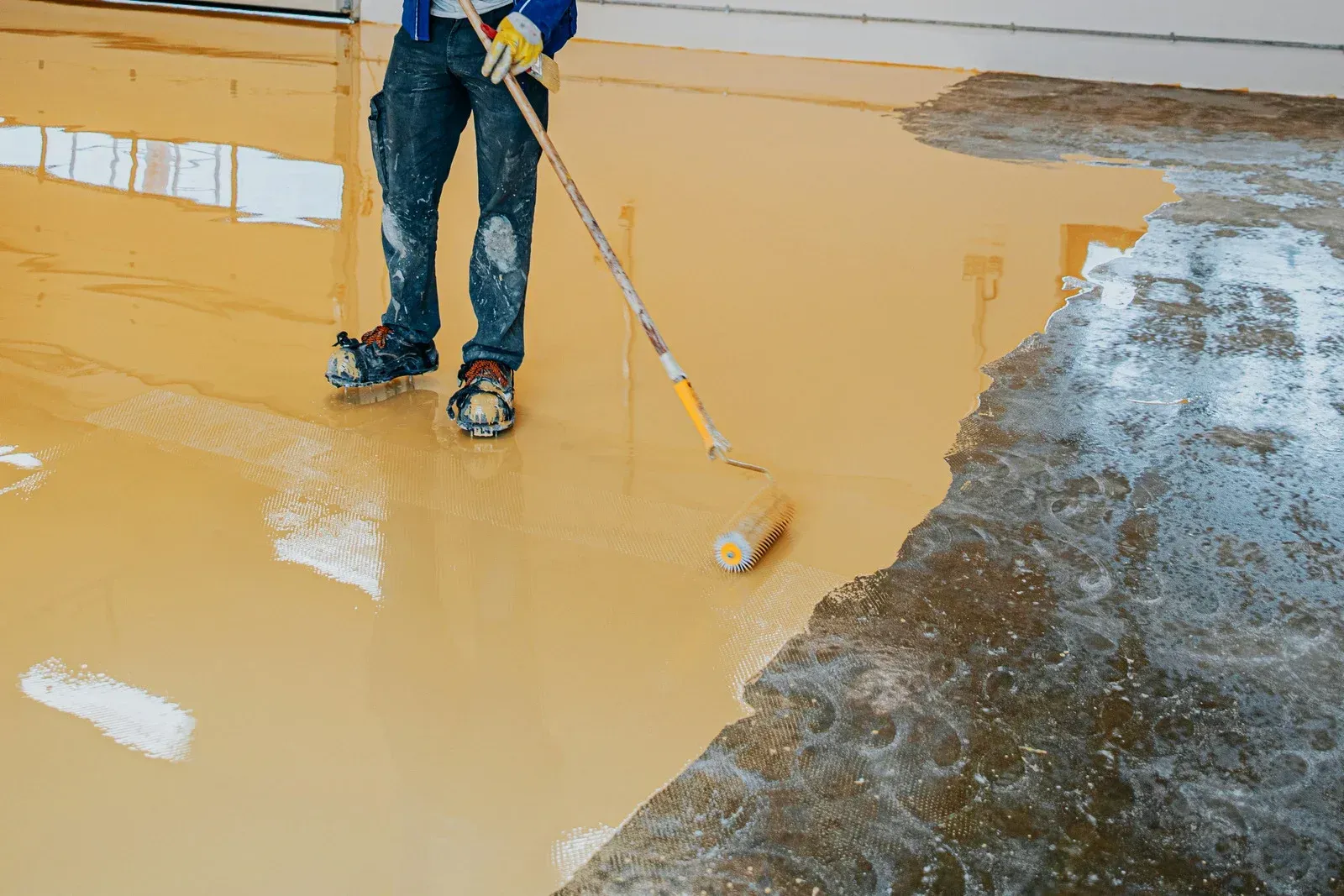 Person spreading yellow coating on a wet floor with a roller; dark rough surface beside it.