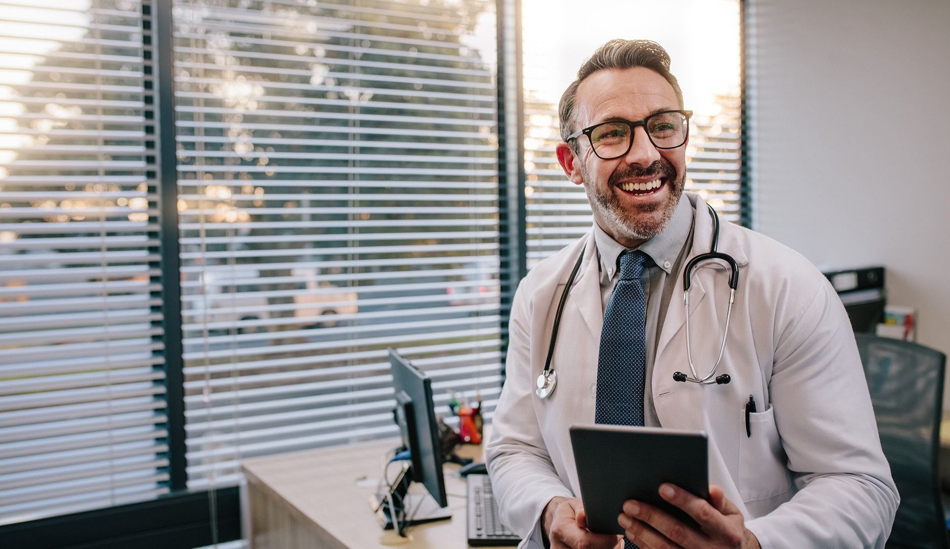 Doctor smiling, holding a tablet, standing in an office with window blinds.