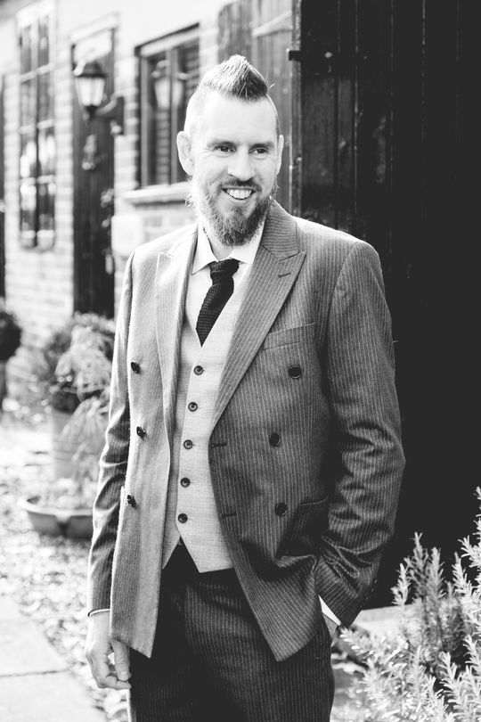 Smiling man in a suit and vest standing outdoors beside a building and potted plants
