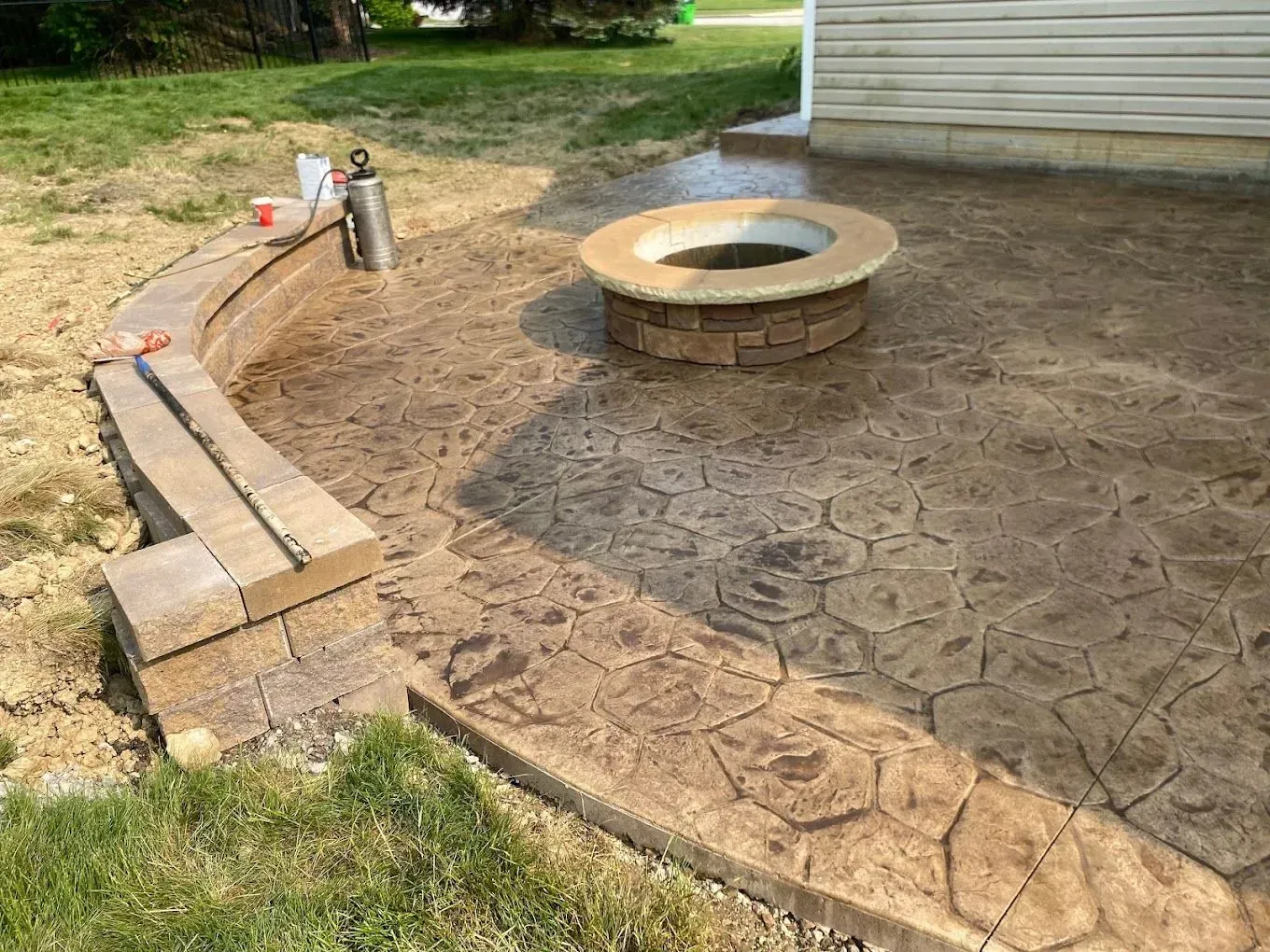 Curved brick patio with fire pit and bench beside a grassy yard and house siding.