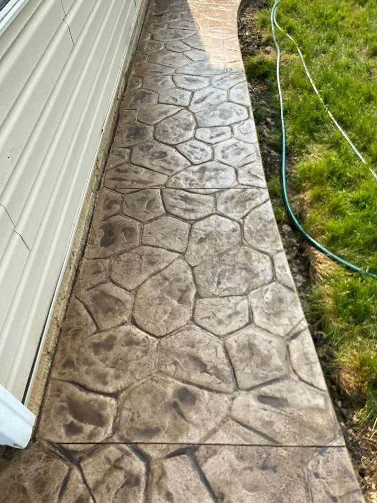 Stamped concrete walkway beside a house, bordered by grass and a green hose.