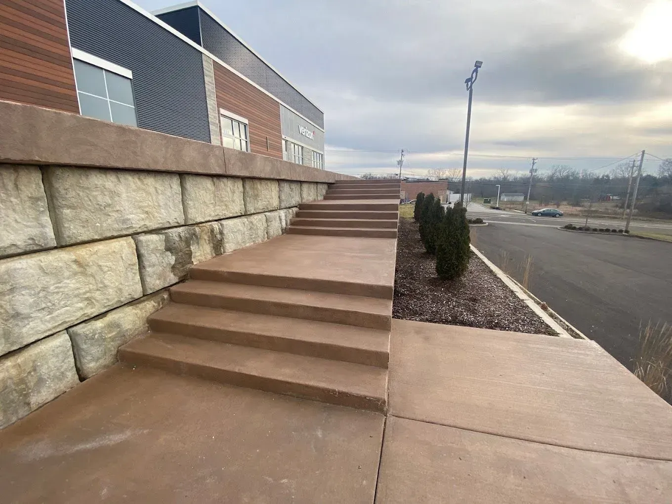 Outdoor concrete stairs beside a stone wall, with a paved road and landscaped embankment on the right.