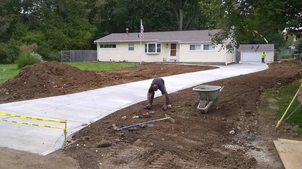 Worker leveling dirt beside a new concrete driveway at a house under construction.