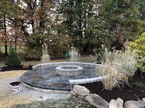 Dry stone fountain basin with a central spout in a landscaped garden, surrounded by trees and shrubs.