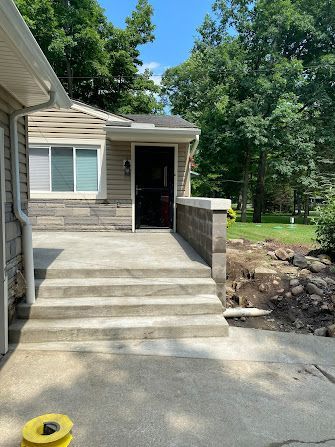 House entry with concrete steps and a ramp-like side wall beside a driveway, surrounded by trees