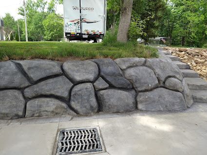 Stone retaining wall beside a sidewalk drain, with grass and a truck in the background.