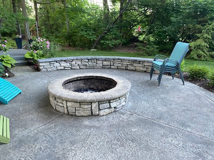 Backyard stone fire pit with curved seating wall and turquoise chairs on a concrete patio, surrounded by trees