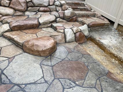 Stone patio with irregular paving and stacked rock steps beside a white fence.