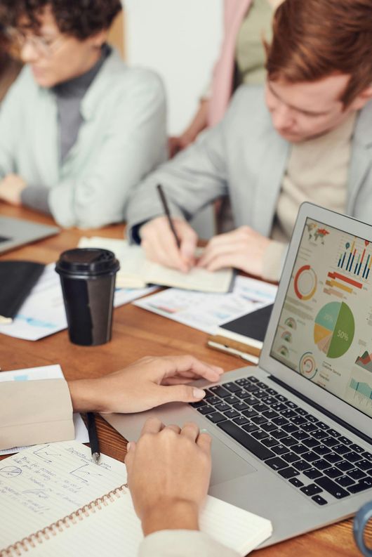 Pessoas em uma reunião: uma trabalhando em um laptop mostrando gráficos, outras fazendo anotações em uma mesa de madeira.
