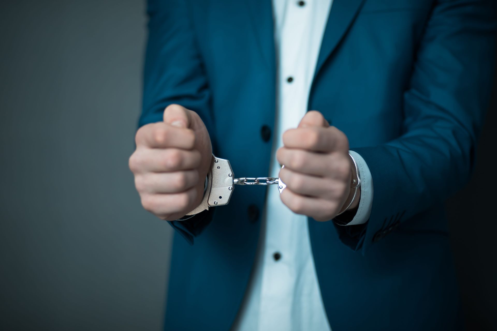A person in a blue suit and white shirt with their wrists handcuffed together against a gray background.