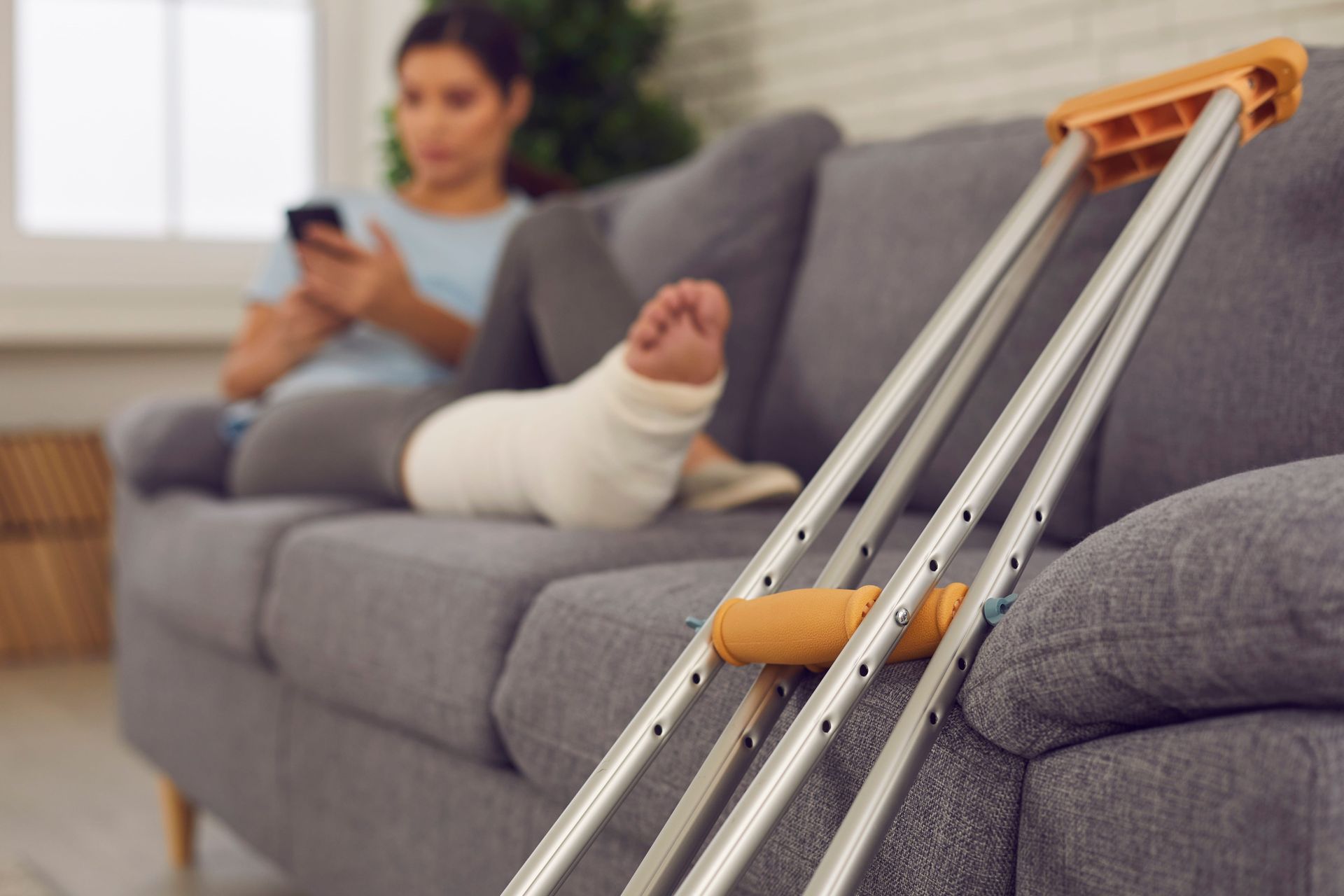 Crutches resting on a gray couch beside a person using a phone, with a leg in a cast in the background