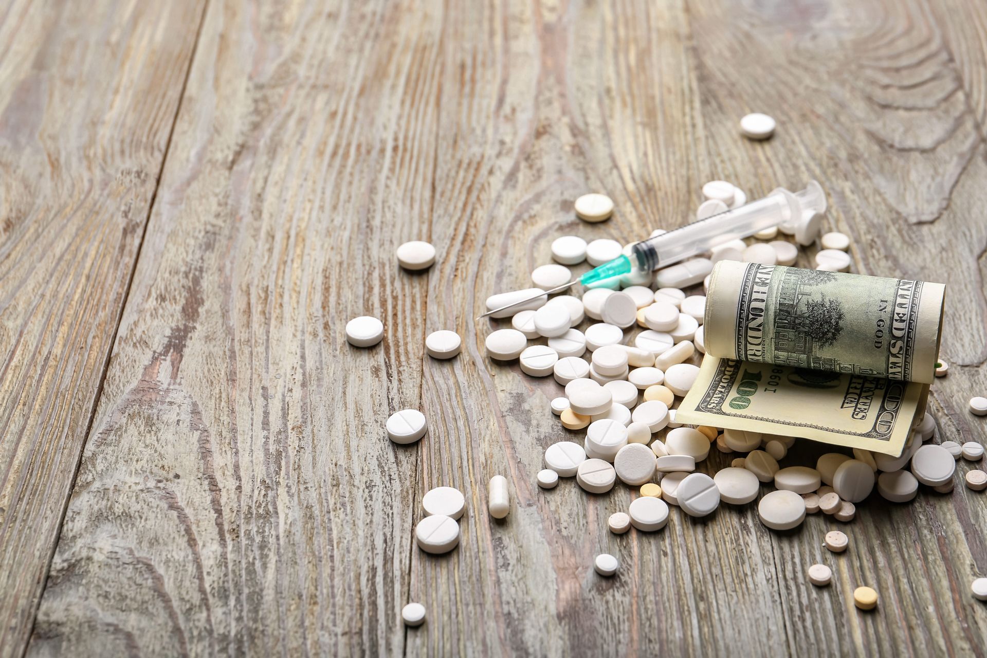 Pills scattered on a wooden surface beside a rolled U.S. dollar bill and a syringe.