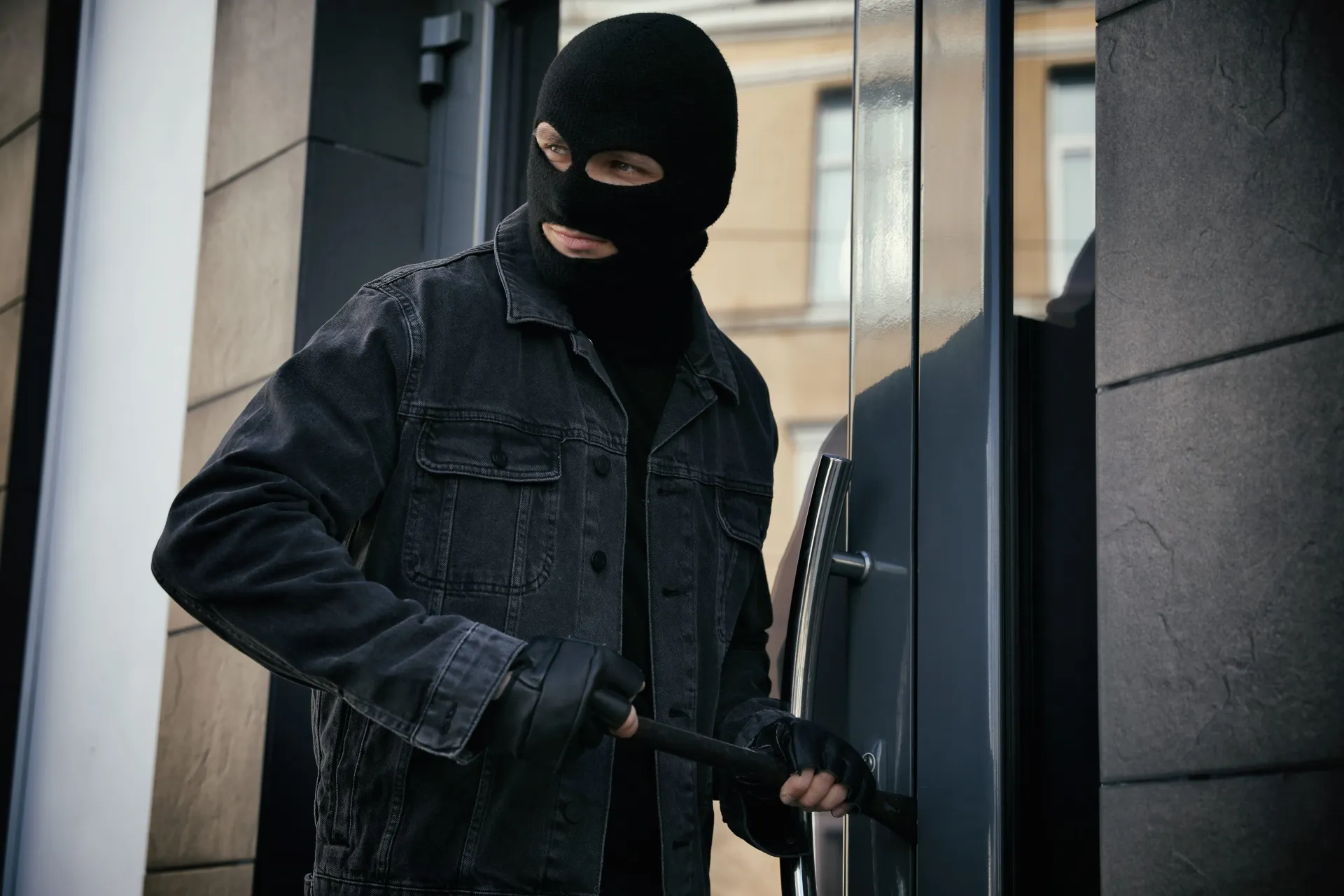 Masked person in dark clothing holding a gun outside a building entrance