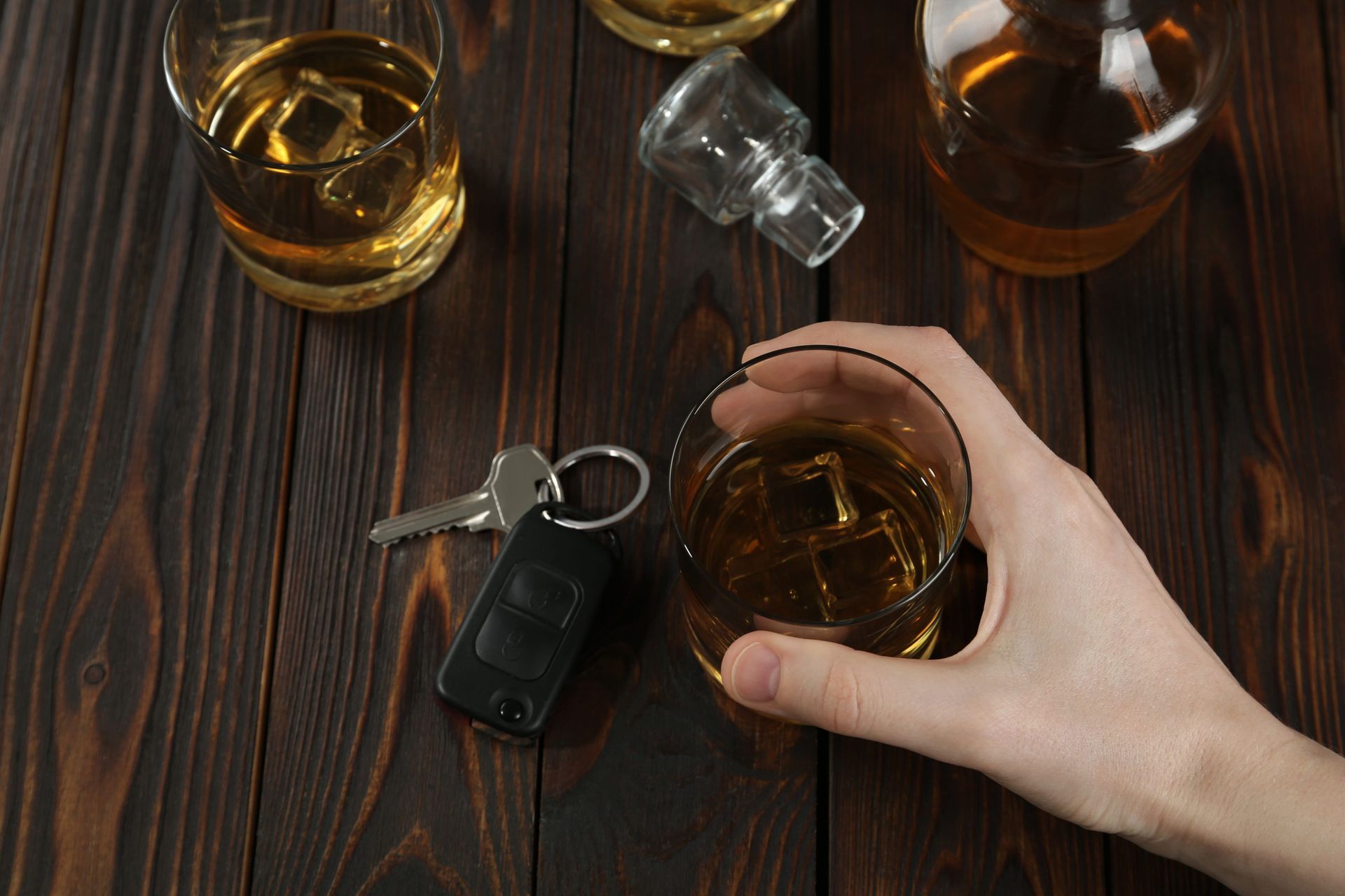 A hand holds a glass of whiskey next to car keys, a bottle, and another glass on a dark wooden table.