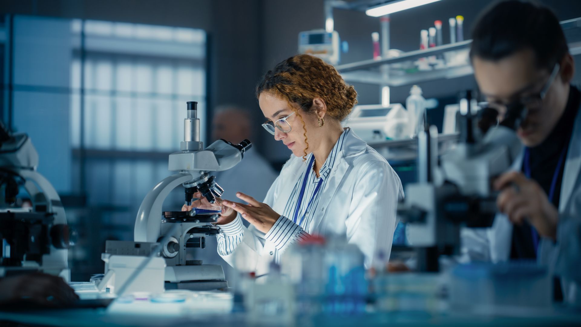 Scientist using a microscope in a laboratory, examining a sample, other scientist present.