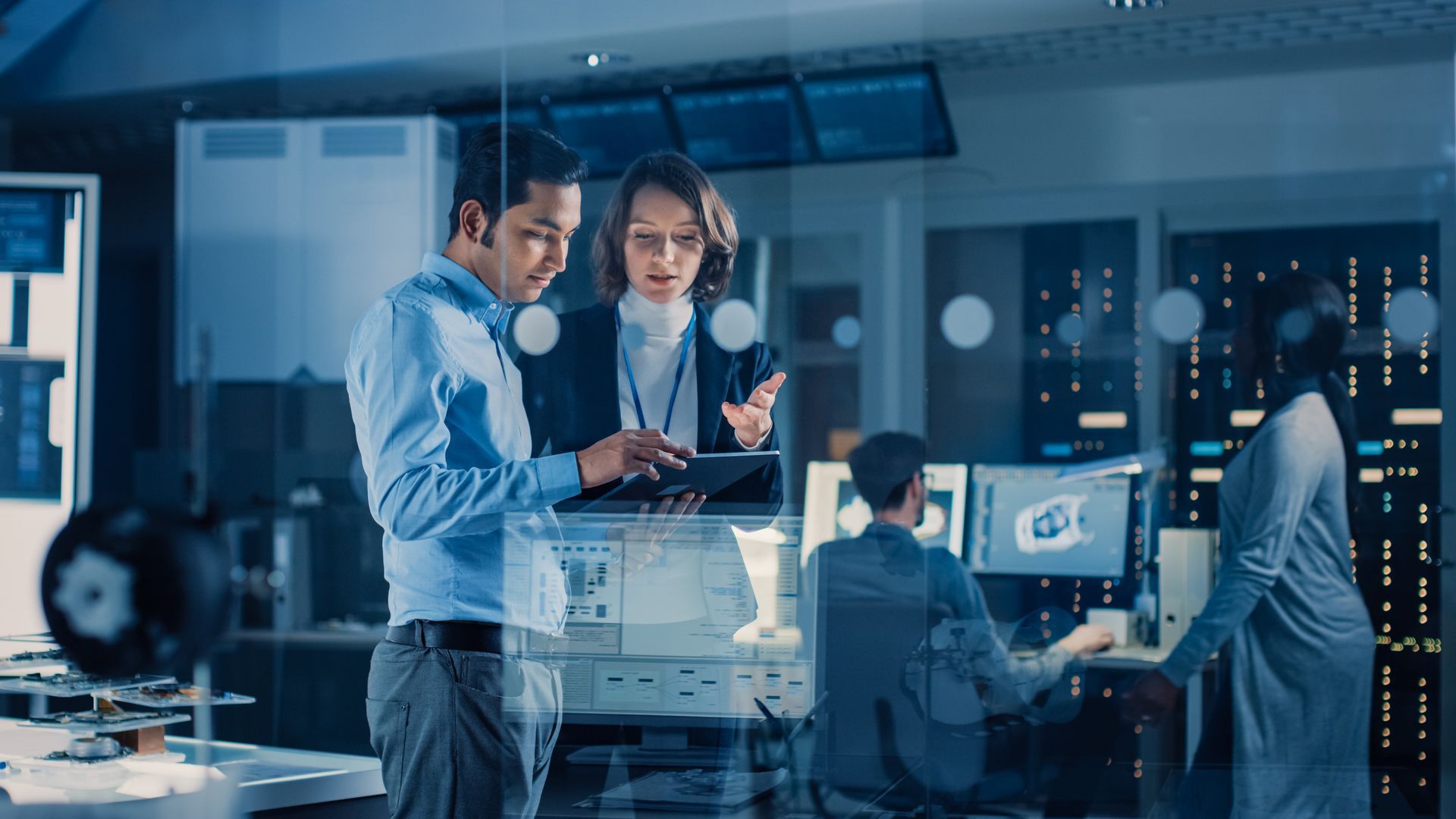 Two people looking at a tablet, consulting in a tech lab. Others work at computers; the room is lit with blue light.