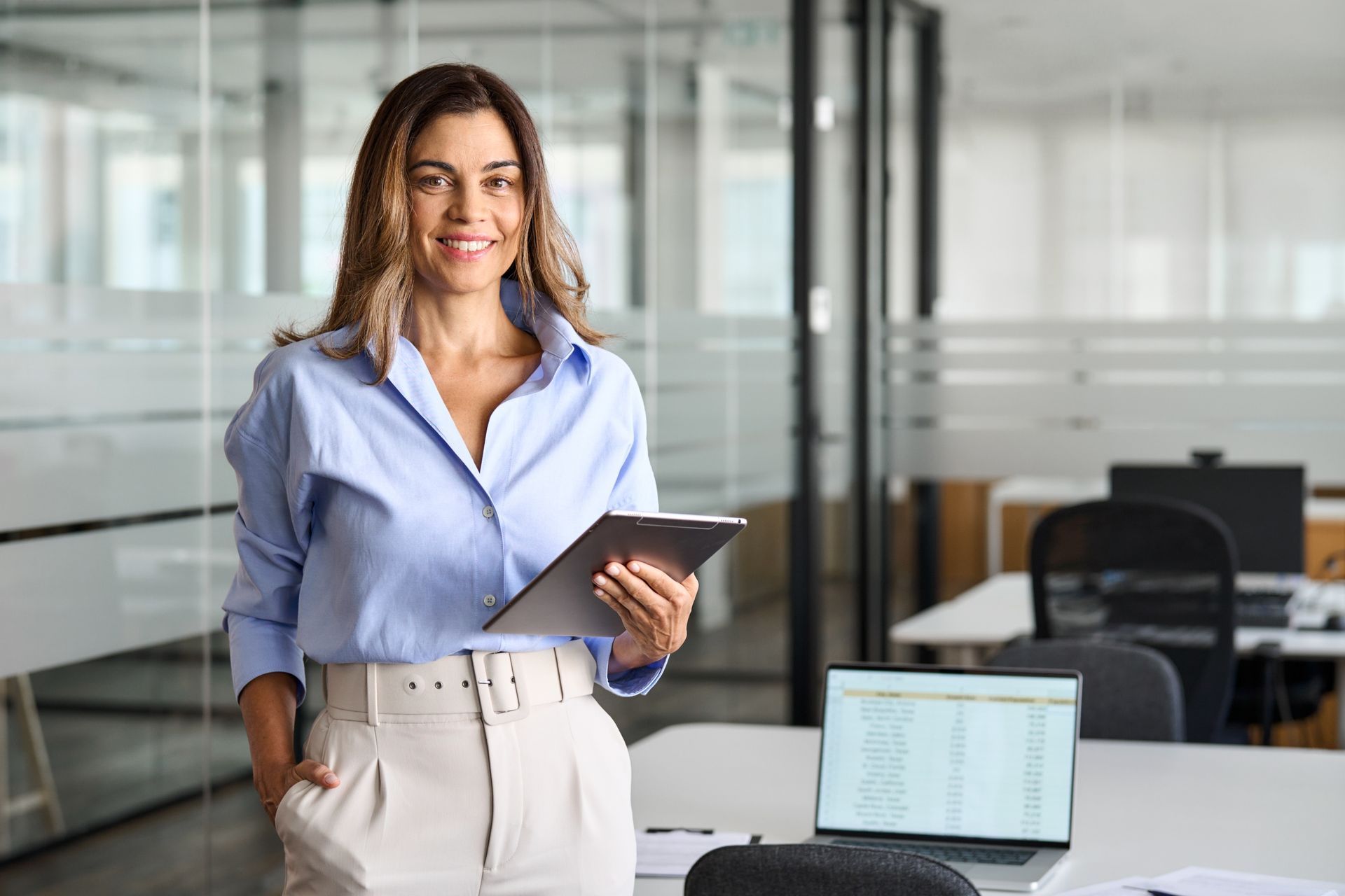 Woman in business attire smiles, holding tablet in office.