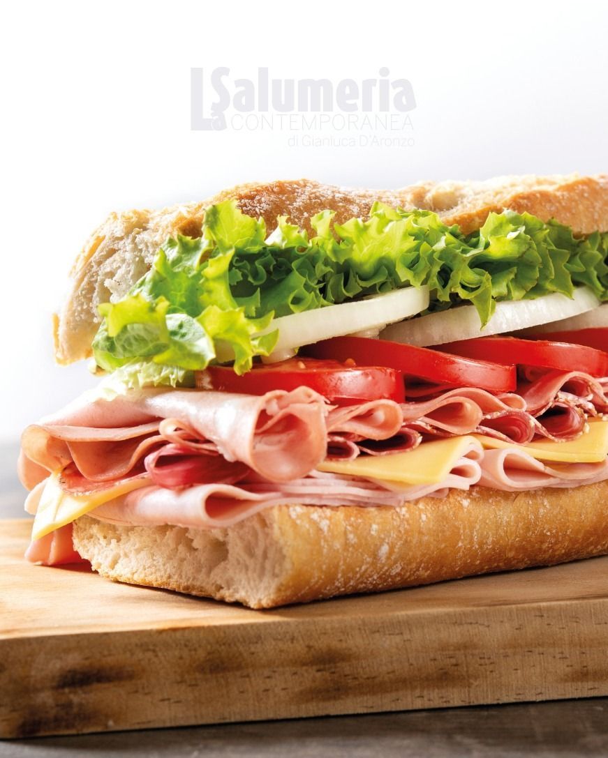 Panino su un tagliere di legno. Contiene fette di prosciutto, formaggio, pomodoro, cipolla e lattuga su una baguette.