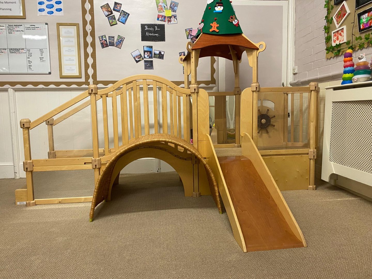 Wooden play structure with a bridge, stairs, and a slide in a classroom.
