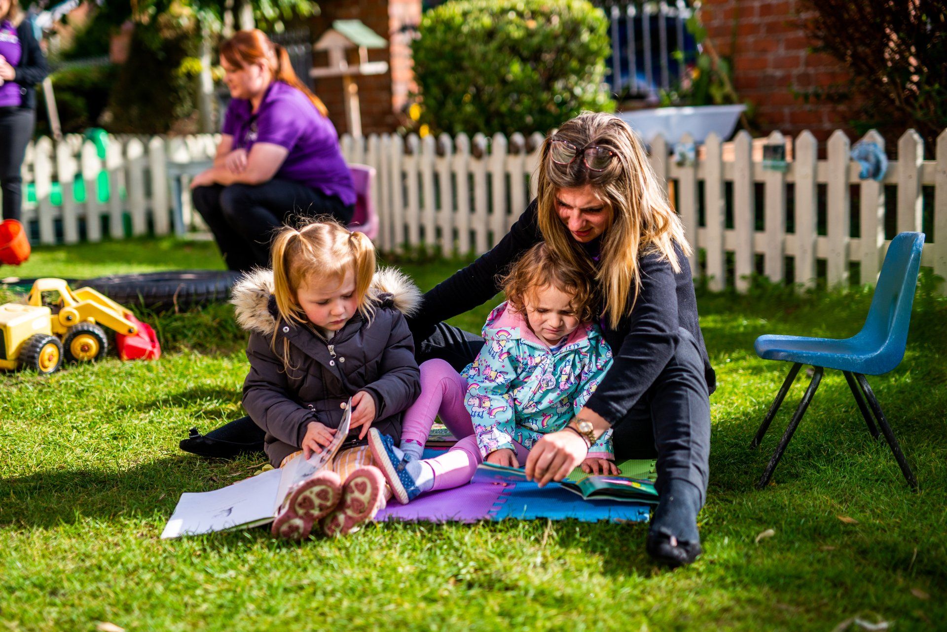 Woman and two children reading on grass; white picket fence and other people in background.