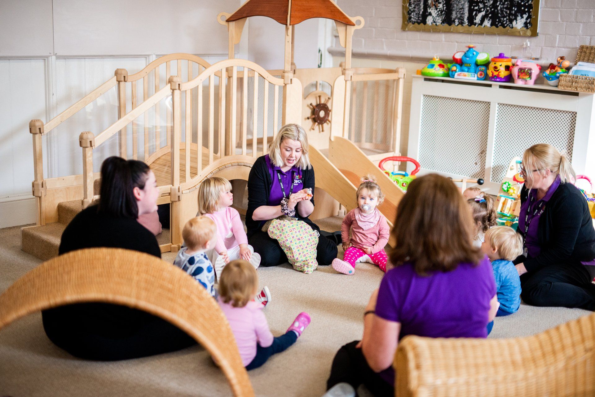 Adults and children sit in a circle in a playroom, some looking at a person holding an item.