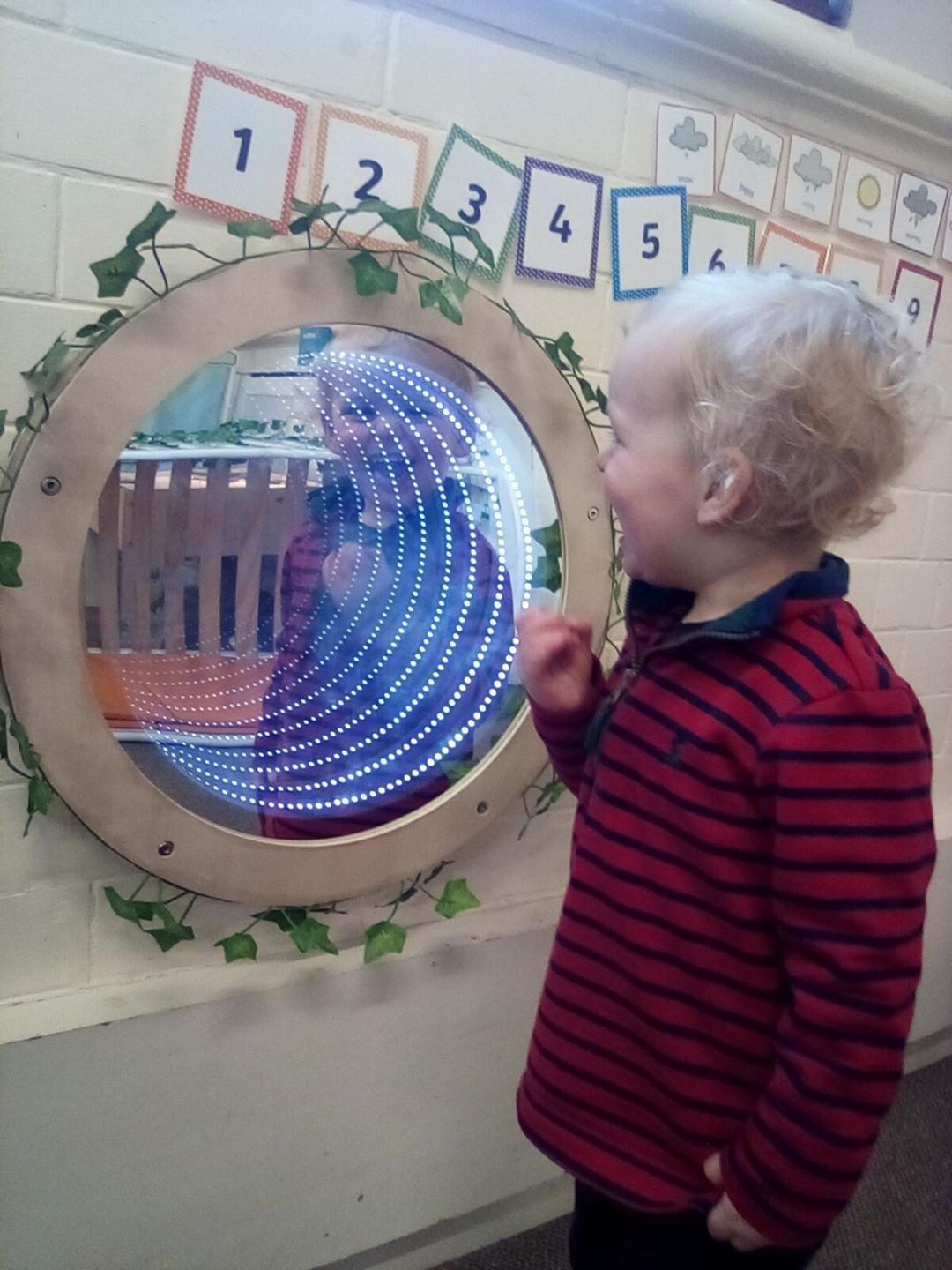 Child looks into a circular mirror with swirling blue lights; ivy decorates the frame.