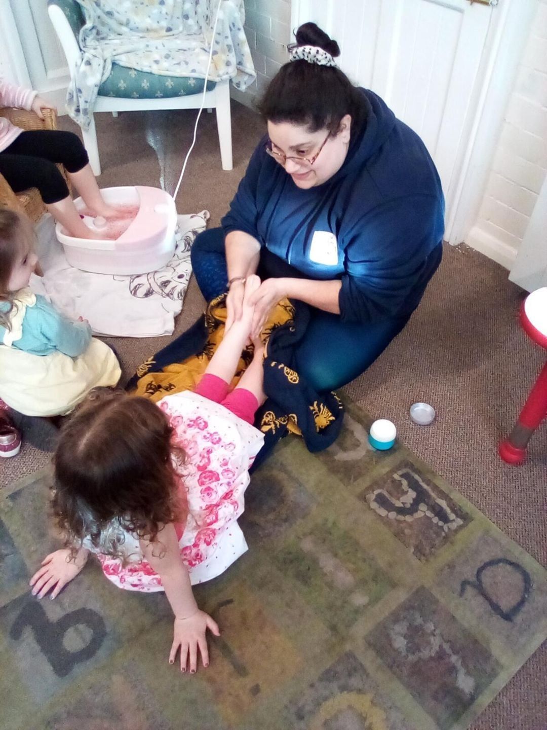 Woman giving child a foot massage on a rug, with other children's feet in a foot bath nearby.