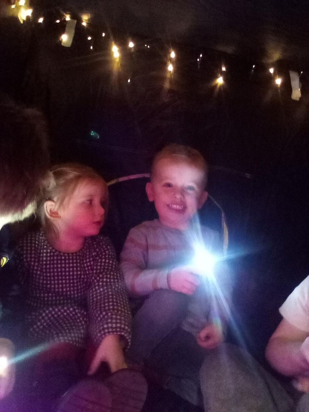 Two children inside a dark tent, one holding a flashlight and smiling, surrounded by string lights.