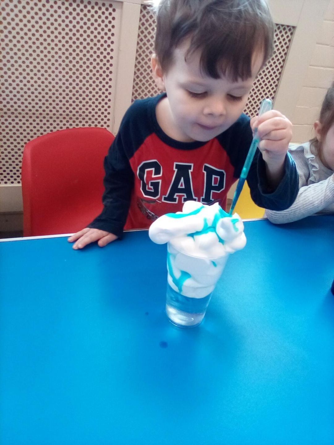 Boy stirring blue-tinted foam in a glass, at a blue table. He wears a red and blue Gap shirt, in a classroom.
